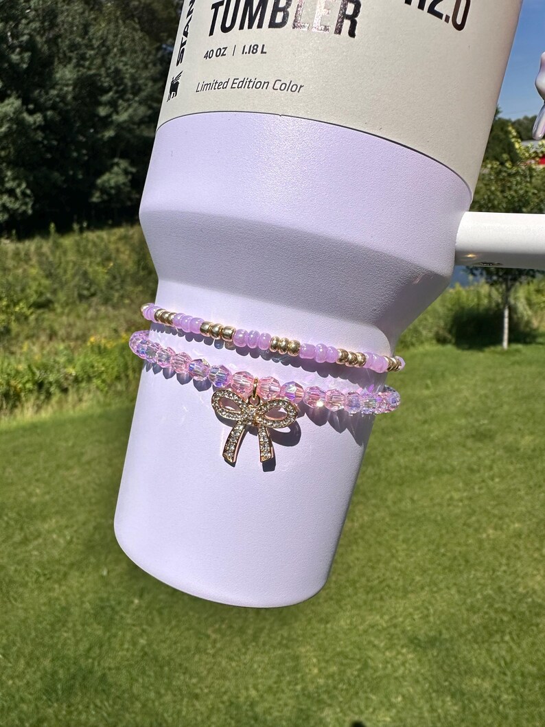 K&ouml;nnte beinhalten: Ein lilafarbener 40 oz Tumbler in limitierter Farbe. Zwei rosafarbene und goldene Perlenarmb&auml;nder mit einem Strass-Schleifenanh&auml;nger sind um den Tumbler gewickelt.