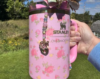 Pode incluir: Um copo Stanley rosa com um padrão floral e uma alça rosa. O copo é adornado com um berloque de contas e um grande laço com detalhes em glitter dourado e rosa. As palavras "LOVESHACKFANCY" e "STANLEY" são visíveis.