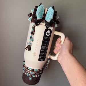 May include: Cream-colored HydroJug water bottle with a handle, adorned with turquoise and brown beaded accents, a decorative bow, and a tassel. The bottle has a capacity of 40oz.