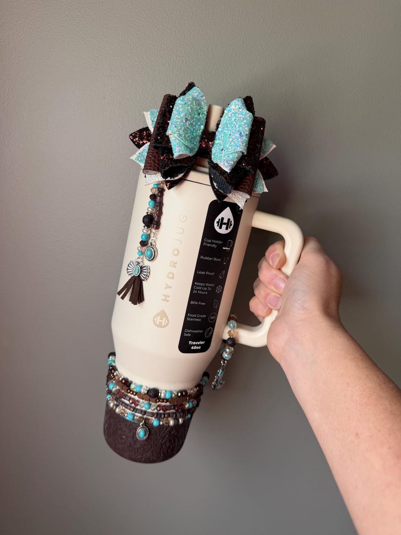 May include: Cream-colored HydroJug water bottle with a handle, adorned with turquoise and brown beaded accents, a decorative bow, and a tassel. The bottle has a capacity of 40oz.