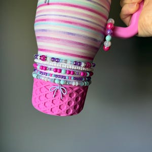 May include: A stainless steel tumbler featuring horizontal stripes in shades of pink, purple, and light blue. The base has a pink silicone honeycomb boot with a light purple bow charm. A stack of colorful beaded bracelets wraps around the tumbler.
