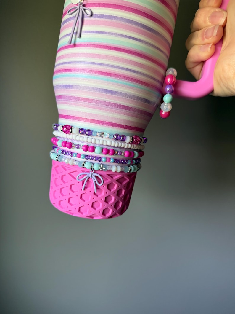 Op de afbeelding: Een roestvrijstalen beker met horizontale strepen in roze, paarse en lichtblauwe tinten. De onderkant heeft een roze siliconen hoes met honingraatpatroon en een lichtpaarse strikbedel. Een stapel kleurrijke kralenarmbanden zit om de beker heen.