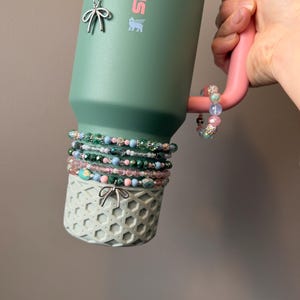 May include: A green Stanley tumbler with a pink handle, adorned with beaded bracelets in shades of pink, green, and blue. Silver charms shaped like bows dangle from the tumbler. The tumbler sits in a light green decorative holder.