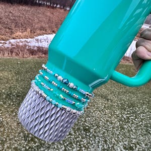 May include: A teal Stanley thermos with a silver base and a handle. The thermos is adorned with multiple beaded bracelets in shades of blue, silver, and teal. The Stanley logo is visible on the side.