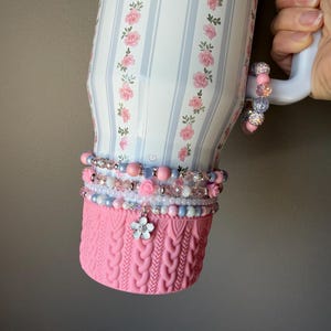 May include: A white tumbler with a floral and striped design, accented with pink and blue beads and a pink knitted sleeve. The tumbler has a handle and is decorated with small flower embellishments.