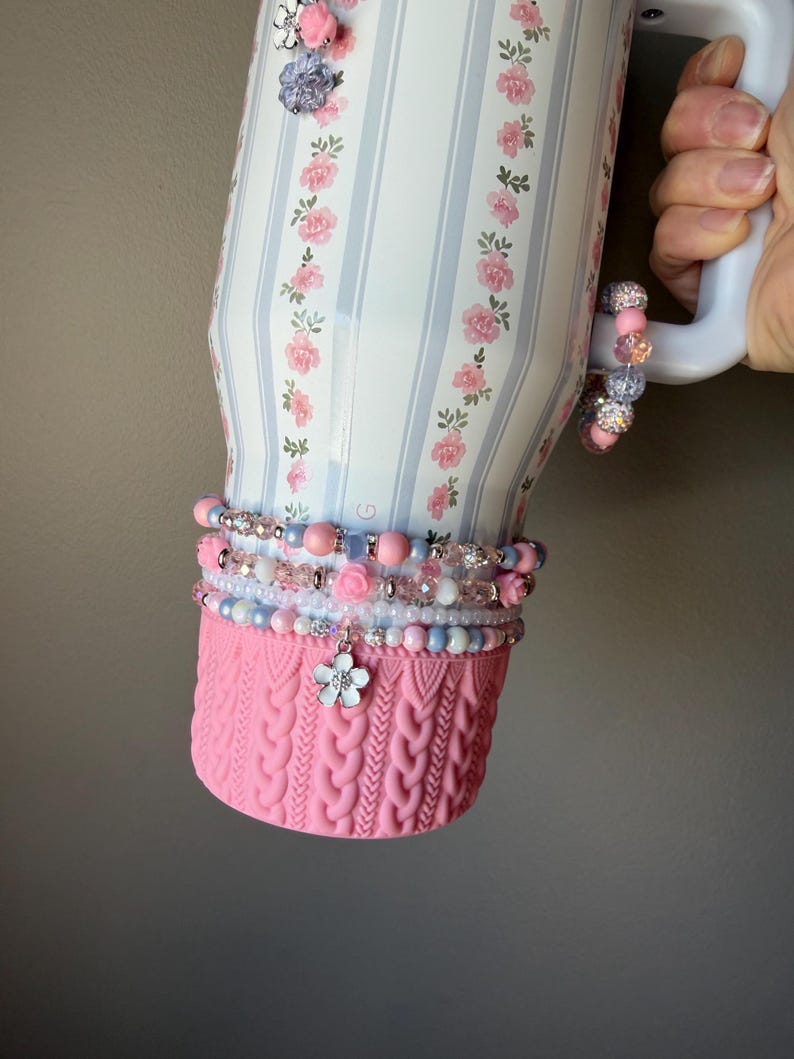 May include: A white tumbler with a floral and striped design, accented with pink and blue beads and a pink knitted sleeve. The tumbler has a handle and is decorated with small flower embellishments.