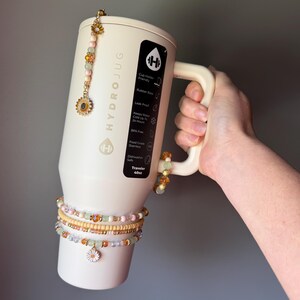 May include: A cream-colored 40oz HydroJug tumbler featuring a black label with white text, adorned with a beaded charm hanging from the lid and three beaded bracelets wrapped around the base, each featuring small flower-shaped metal charms.