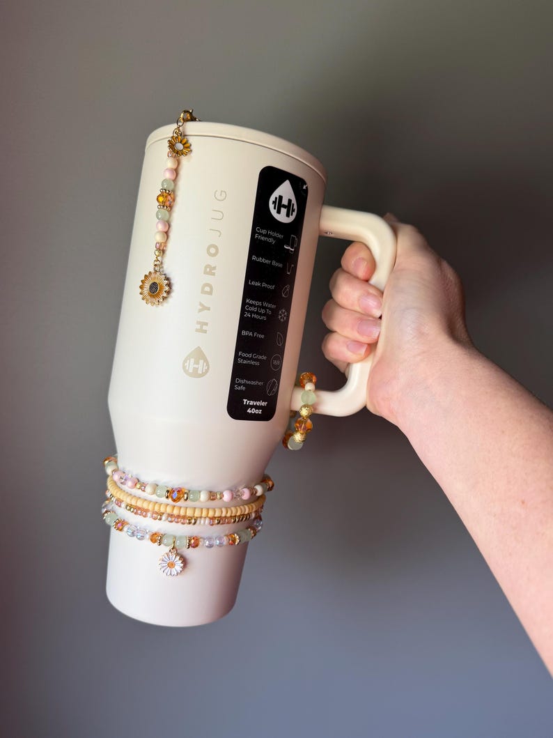 May include: A cream-colored 40oz HydroJug tumbler featuring a black label with white text, adorned with a beaded charm hanging from the lid and three beaded bracelets wrapped around the base, each featuring small flower-shaped metal charms.