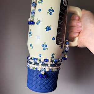 Op de afbeelding: Een cr&egrave;mekleurige 32oz roestvrijstalen beker met een bosbessenpatroon, een handvat en een donkerblauwe siliconen basis met een gevlochten textuur. De beker is versierd met meerdere kralenbedels in blauwe, witte en groene tinten, inclusief kleine bloem- en besaccenten.