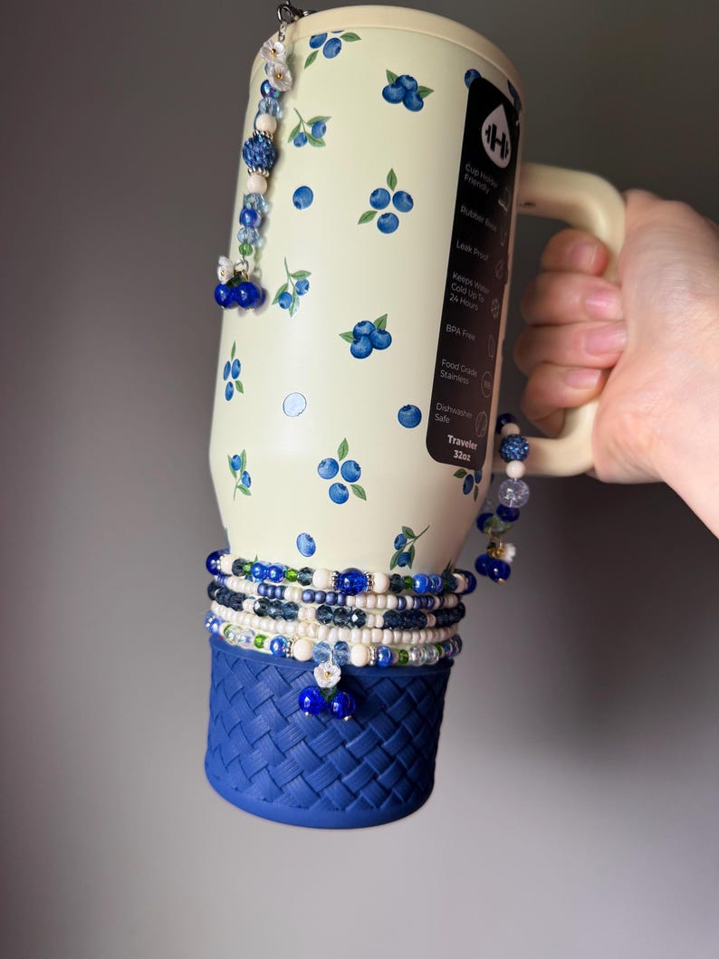 Op de afbeelding: Een cr&egrave;mekleurige 32oz roestvrijstalen beker met een bosbessenpatroon, een handvat en een donkerblauwe siliconen basis met een gevlochten textuur. De beker is versierd met meerdere kralenbedels in blauwe, witte en groene tinten, inclusief kleine bloem- en besaccenten.