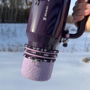May include: A purple Stanley + Starbucks tumbler with a handle, adorned with beaded bracelets and silver bow charms. The bottom of the tumbler has a light purple, textured sleeve. The background is a snowy landscape.