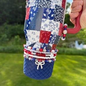Puede incluir: Un vaso t&eacute;rmico grande con un patr&oacute;n de patchwork en rojo, blanco y azul con dise&ntilde;os florales y geom&eacute;tricos. Tiene un asa roja y una base azul texturizada. El vaso est&aacute; decorado con m&uacute;ltiples dijes de cuentas y un dije de lazo plateado.