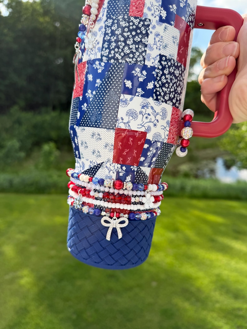 Puede incluir: Un vaso t&eacute;rmico grande con un patr&oacute;n de patchwork en rojo, blanco y azul con dise&ntilde;os florales y geom&eacute;tricos. Tiene un asa roja y una base azul texturizada. El vaso est&aacute; decorado con m&uacute;ltiples dijes de cuentas y un dije de lazo plateado.