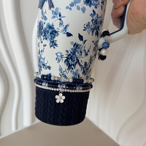 May include: A white tumbler with a blue floral design, a navy blue knitted sleeve, and beaded bracelets. A blue bow and a flower charm are attached. The tumbler is held by a person.