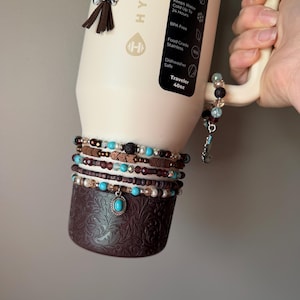 May include: A cream-colored insulated water bottle with a brown base and handle, adorned with turquoise and brown beaded bracelets and charms. The bottle has a black label with text, including "Traveler 40oz."