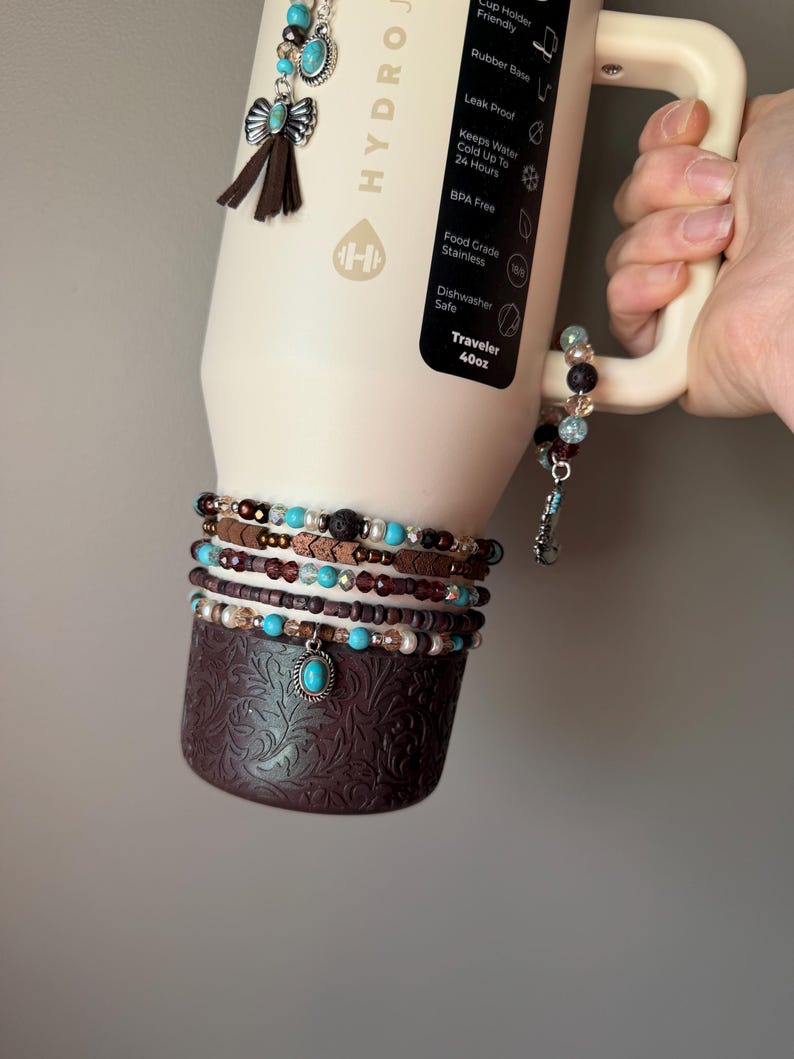 May include: A cream-colored insulated water bottle with a brown base and handle, adorned with turquoise and brown beaded bracelets and charms. The bottle has a black label with text, including "Traveler 40oz."