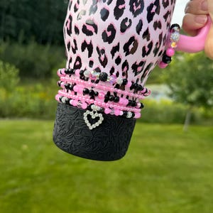 May include: A pink tumbler with a black leopard print pattern featuring a pink handle and a black base with an embossed floral texture. The tumbler is adorned with a stack of beaded bracelets in shades of pink and black, featuring a heart-shaped charm.