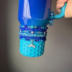 May include: A vibrant blue tumbler with a turquoise base, adorned with multiple beaded bracelets in shades of blue and silver. A silver charm in the shape of a whale tail hangs from the bracelets. A silver wave charm is attached to the top of the tumbler.