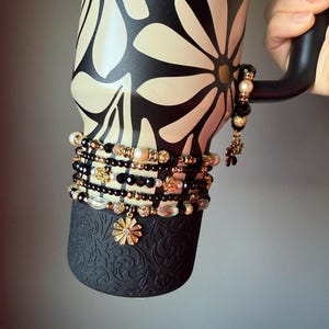 May include: A black and cream floral-patterned tumbler with a black handle. The tumbler is adorned with multiple beaded bracelets in black, gold, and clear accents. A gold flower charm hangs from one of the bracelets.