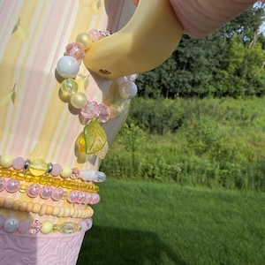 May include: A beaded charm featuring translucent pink, yellow, and white beads with a green leaf pendant, attached to the handle of a pale yellow and pink striped tumbler. The tumbler has a textured pink base and a lemon-patterned design.