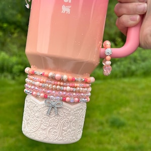 May include: A pink and peach ombre Stanley tumbler with a decorative white base featuring an ornate floral design. The tumbler is adorned with multiple beaded bracelets in shades of pink, peach, and silver. A silver bow charm is attached to the base.