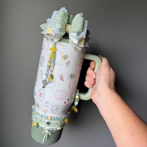 May include: A white travel mug with a green handle and base, adorned with a large bow and beaded accents. The mug features a botanical design with the words "La Dolce Vita" and is embellished with a charm. The mug is held by a person.