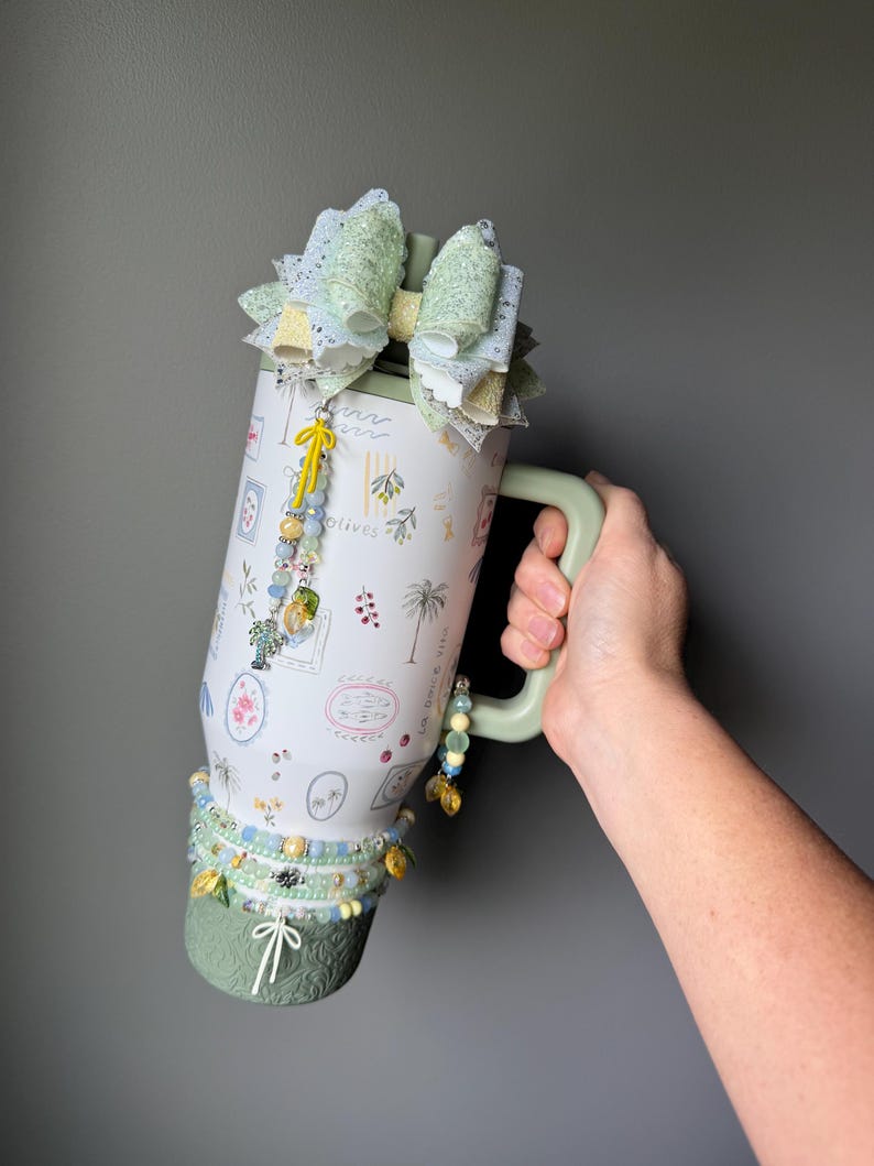 May include: A white travel mug with a green handle and base, adorned with a large bow and beaded accents. The mug features a botanical design with the words "La Dolce Vita" and is embellished with a charm. The mug is held by a person.