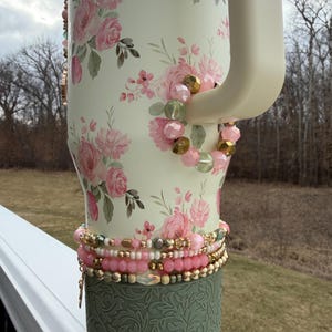 May include: A cream-coloured tumbler decorated with pink floral patterns and a green base with an embossed design. The tumbler is accessorised with beaded bracelets in shades of pink, gold, and green.
