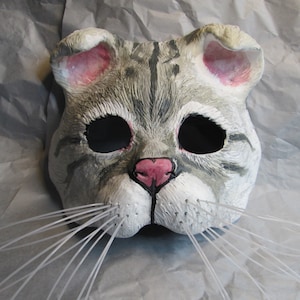 Pode incluir: Uma máscara de gato cinza e branco com orelhas cor-de-rosa e um nariz preto. A máscara tem bigodes brancos e buracos pretos para os olhos.