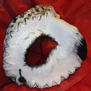 May include: A white and black owl mask with a large eye opening. The mask has a textured surface with black accents and a black string attached. The mask is set against a red background.