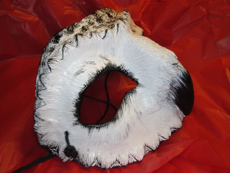 May include: A white and black owl mask with a large eye opening. The mask has a textured surface with black accents and a black string attached. The mask is set against a red background.