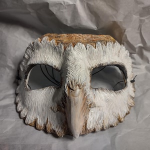 May include: A handcrafted owl mask with a textured, feathered appearance. The mask features a white and brown color scheme, with detailed carving around the eyes and beak. Black string is attached for wearing.