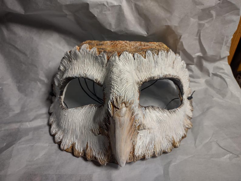 May include: A handcrafted owl mask with a textured, feathered appearance. The mask features a white and brown color scheme, with detailed carving around the eyes and beak. Black string is attached for wearing.