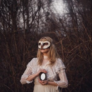 May include: A person wearing a handmade owl mask and a white lace dress, holding a small oval mirror. The mask is primarily cream and brown, with detailed feather-like textures. The dress features delicate lace sleeves and a high neckline.
