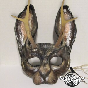 May include: A handcrafted deer mask with large ears and attached antlers. The mask is primarily brown with white and tan accents, and features eye openings and a nose. The mask is designed for costume or theatrical use.