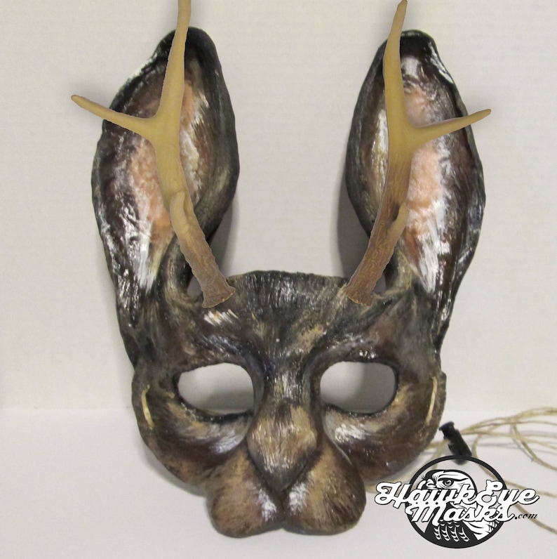 May include: A handcrafted deer mask with large ears and attached antlers. The mask is primarily brown with white and tan accents, and features eye openings and a nose. The mask is designed for costume or theatrical use.
