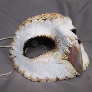 May include: A handcrafted owl mask with a white, textured surface, detailed with brown and tan accents. The mask features a beak with red and gray paint, and a string for wearing. The eye opening is black.