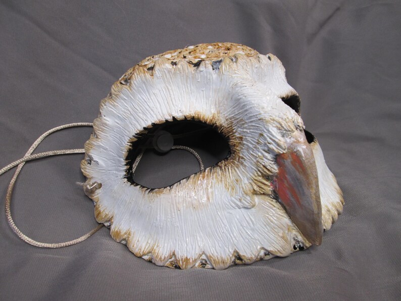May include: A handcrafted owl mask with a white, textured surface, detailed with brown and tan accents. The mask features a beak with red and gray paint, and a string for wearing. The eye opening is black.