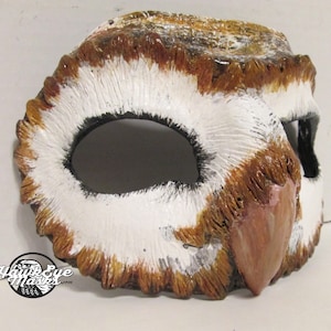 May include: A handcrafted owl mask with a detailed design. The mask features a white, brown, and black color scheme, with textured feathers and a prominent beak. The eye openings are large and dark, and the mask appears to be made of a durable material.