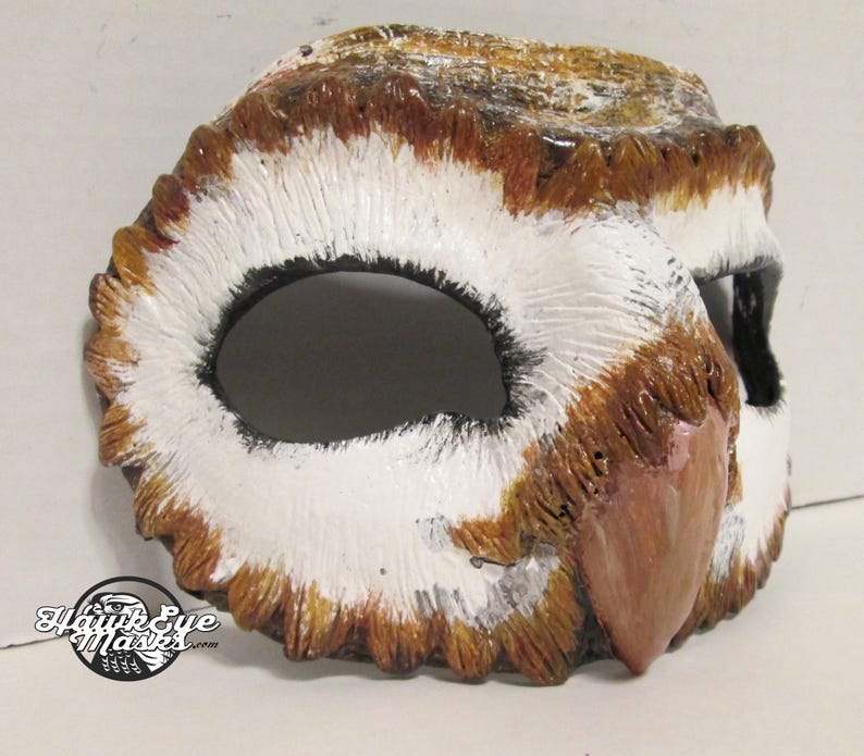 May include: A handcrafted owl mask with a detailed design. The mask features a white, brown, and black color scheme, with textured feathers and a prominent beak. The eye openings are large and dark, and the mask appears to be made of a durable material.