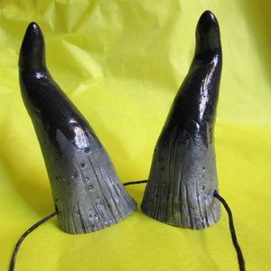 May include: A pair of black and gray devil horns with a textured surface. The horns are painted black at the tips, fading to gray at the base. Each horn has a small hole with a black string attached.