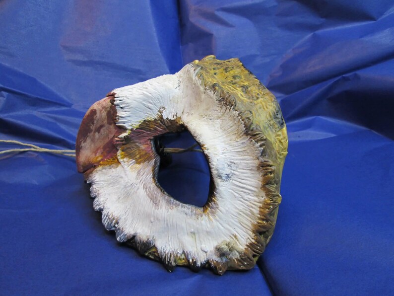 May include: A handmade, avian-themed mask with a central eye opening. The mask features a combination of white, brown, and yellow paint, with a textured surface. The mask is designed to be worn and is likely for costume or decorative purposes.