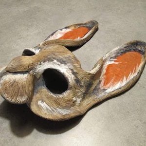 May include: A handcrafted, decorative rabbit mask. The mask is primarily brown with white and black accents, and features orange-painted inner ears. The mask has a large eye opening and is designed to be worn.