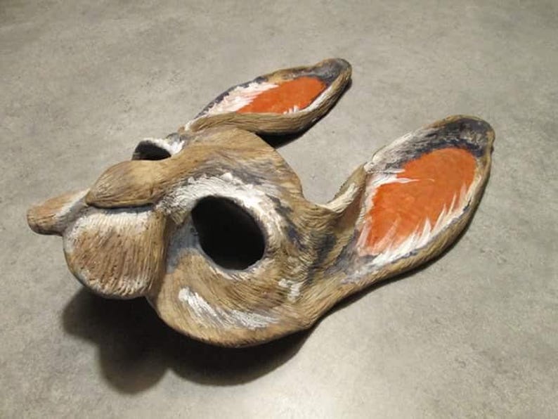 May include: A handcrafted, decorative rabbit mask. The mask is primarily brown with white and black accents, and features orange-painted inner ears. The mask has a large eye opening and is designed to be worn.
