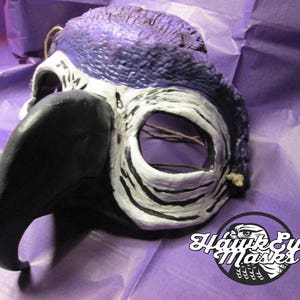 May include: A purple, black, and white bird mask with a long beak. The mask is made of papier-mâché and has a textured surface. The mask is for a masquerade ball or costume party.  The mask is handmade and has a unique design.  The mask is for sale on Etsy.  Hawk Eye Masks.com