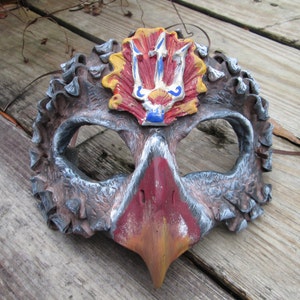 Könnte beinhalten: Eine handbemalte Theatermaske mit einem vogelartigen Design. Die Maske hat eine braune und graue strukturierte Oberfläche mit einem roten und orangefarbenen Schnabel. Die Oberseite der Maske hat ein gelbes, rotes und blaues Emblem. Die Augenlöcher sind groß und oval.