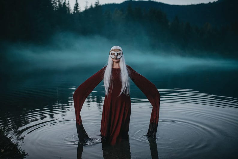 May include: A person stands in water, wearing a burgundy dress with long, flowing sleeves. They wear a decorative owl mask and have long, white hair. The background features a misty lake and dark forest.