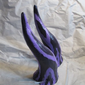May include: A pair of black and purple horns with a textured surface. The horns are made of a lightweight material and are designed to be worn as a costume accessory.