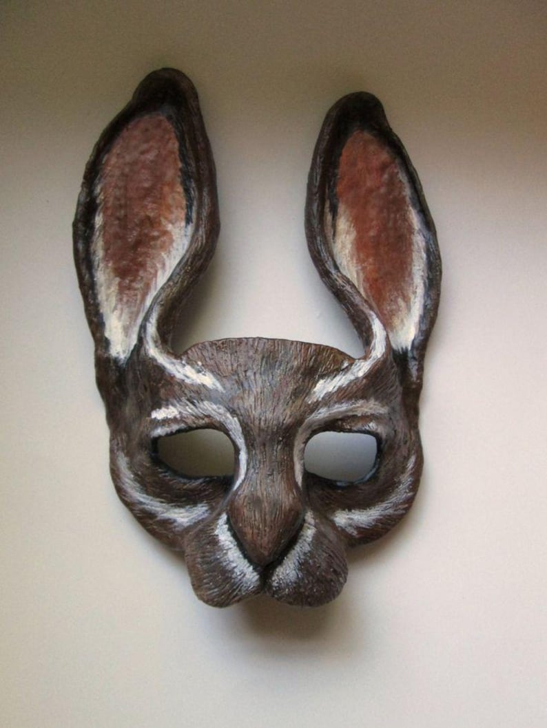 May include: A handcrafted rabbit mask with long, upright ears. The mask is primarily brown with white and reddish-brown accents inside the ears. The eye holes are oval-shaped, and the mask has a textured appearance, suggesting a natural material.