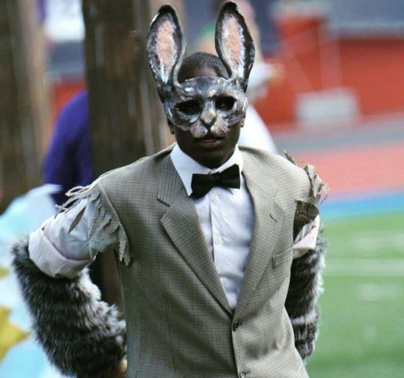 May include: A person in a gray suit and a rabbit mask with long ears. The suit has torn sleeves and faux fur cuffs. A black bow tie is worn with a white collared shirt. The background is blurred, suggesting an outdoor event.